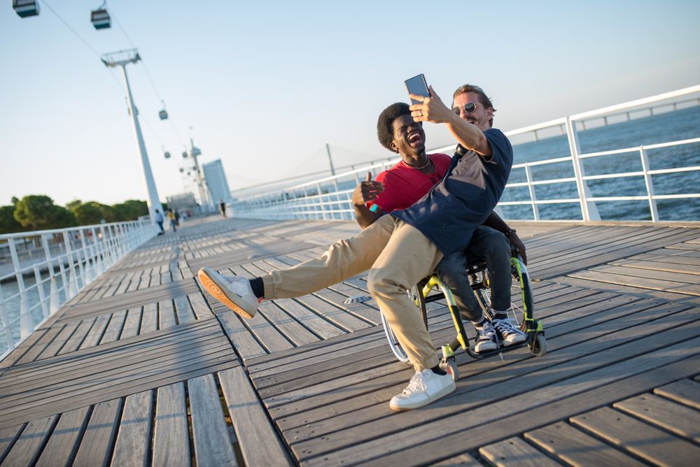 Dos amigos se hacen un selfie en un paseo marítimo; uno va en silla de ruedas.